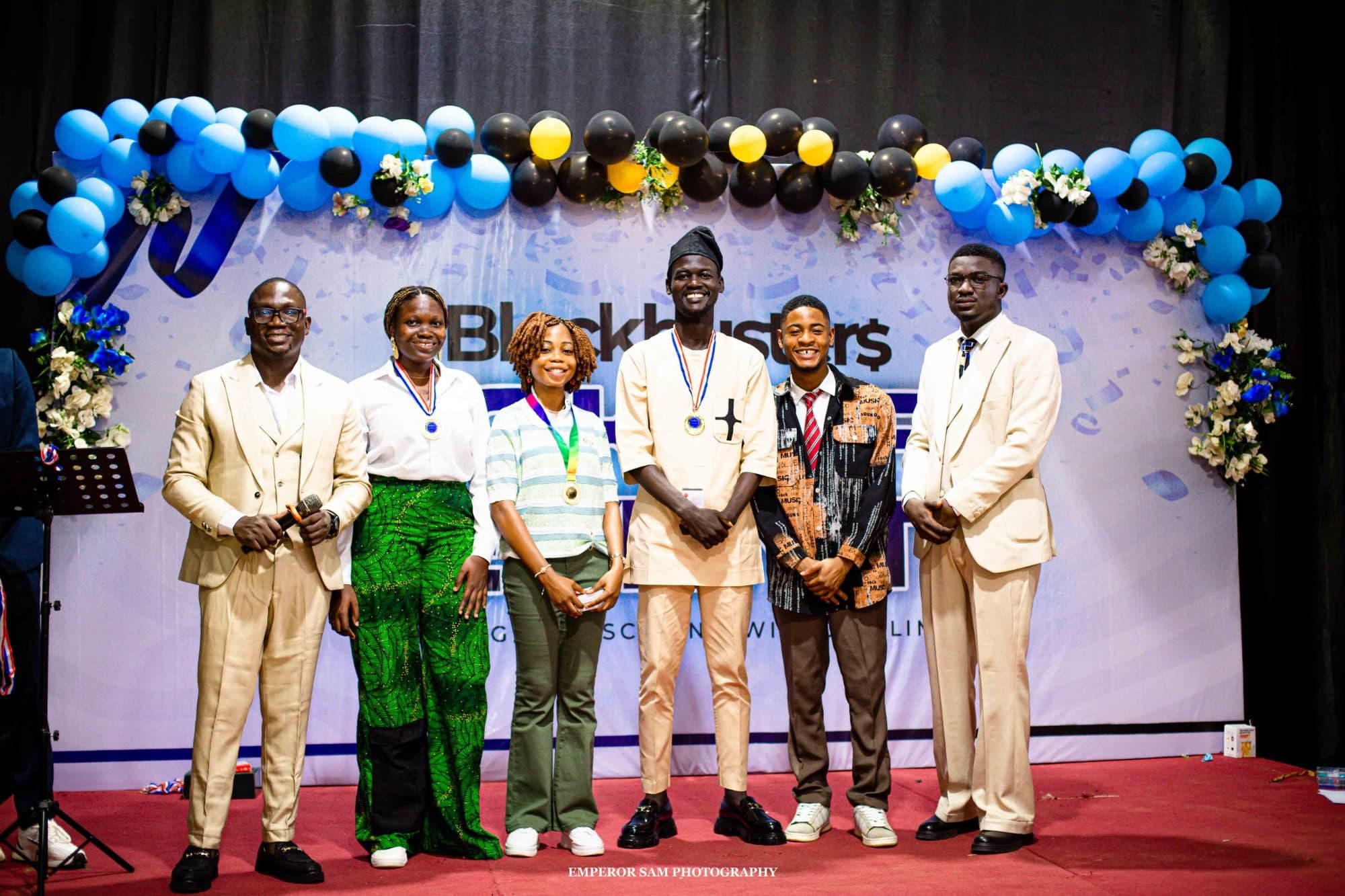 Award winners wearing medals stand on stage with hosts in front of a balloon arch