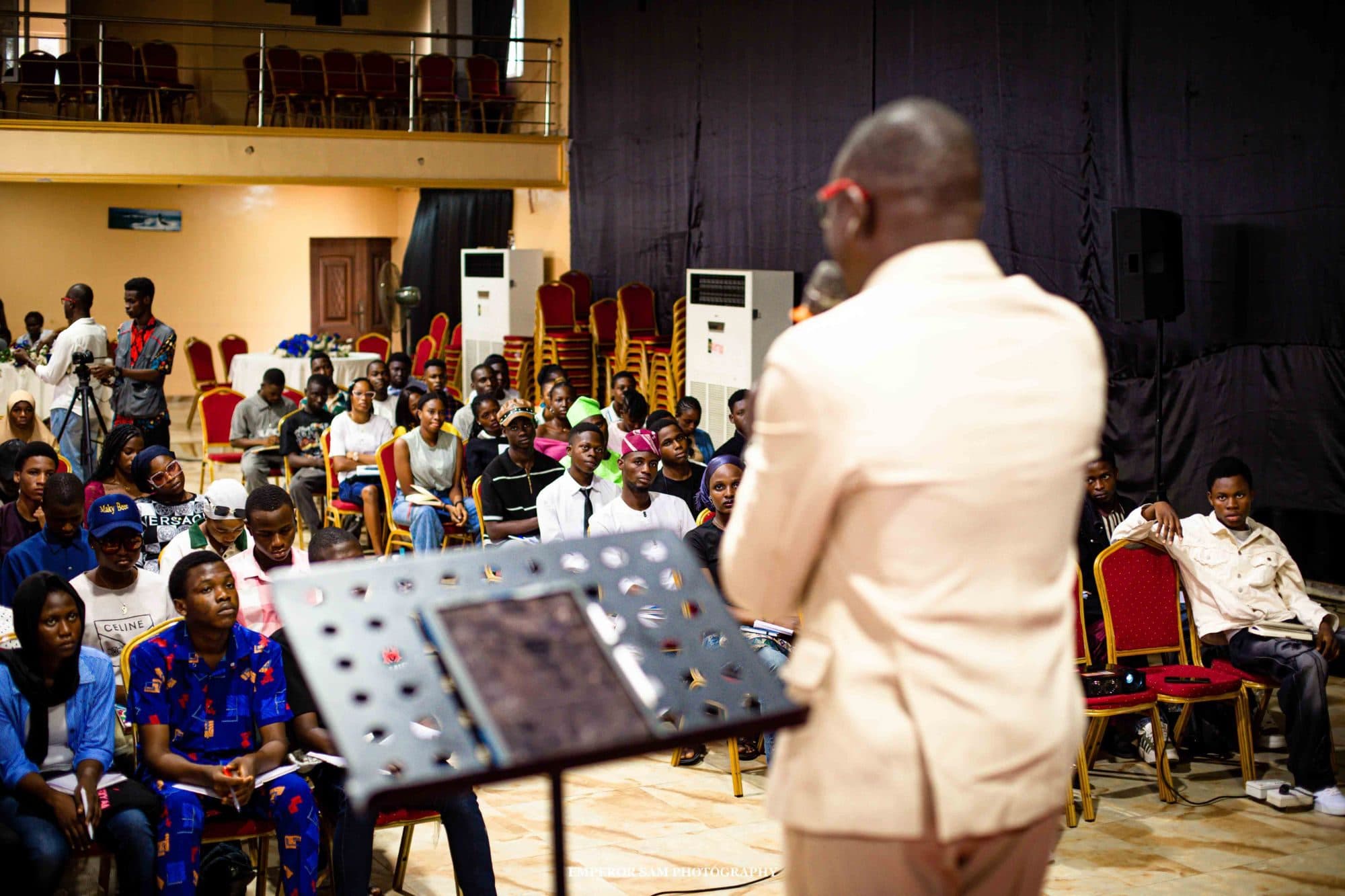 A speaker in a cream suit addresses a seated audience of young members taking notes